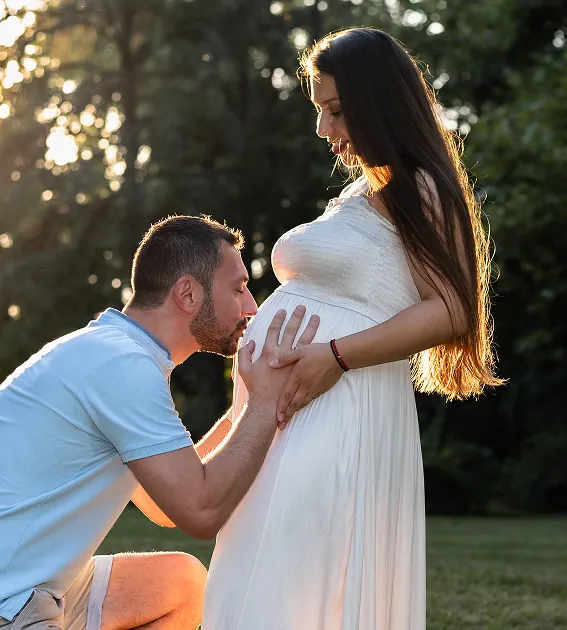 Partner ljubi trudnički stomak žene u beloj haljini na suncu